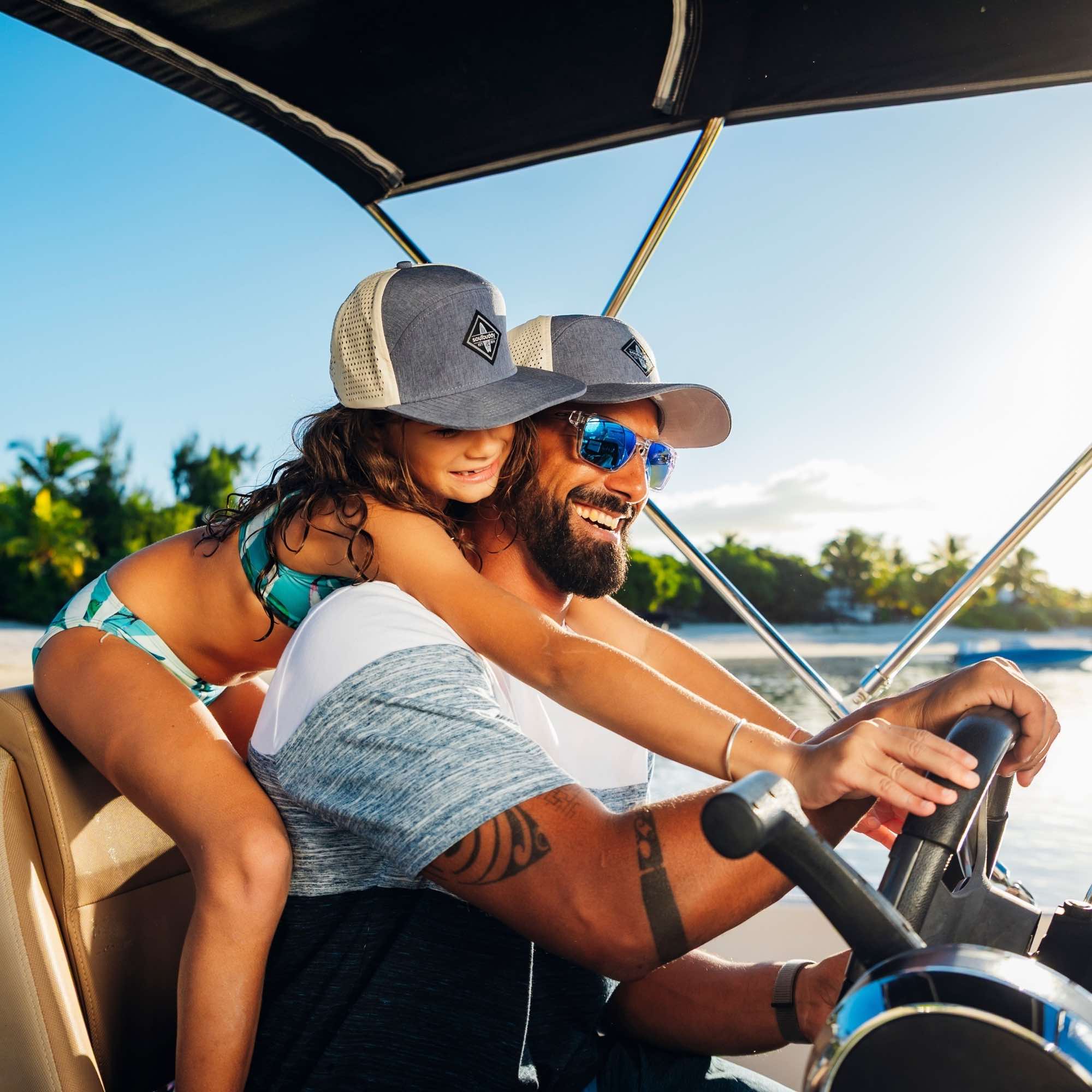 Vater und Tochter tragen graue Snapback Caps im Partnerlook und genießen gemeinsamen Bootsausflug am Meer bei sommerlichem Wetter