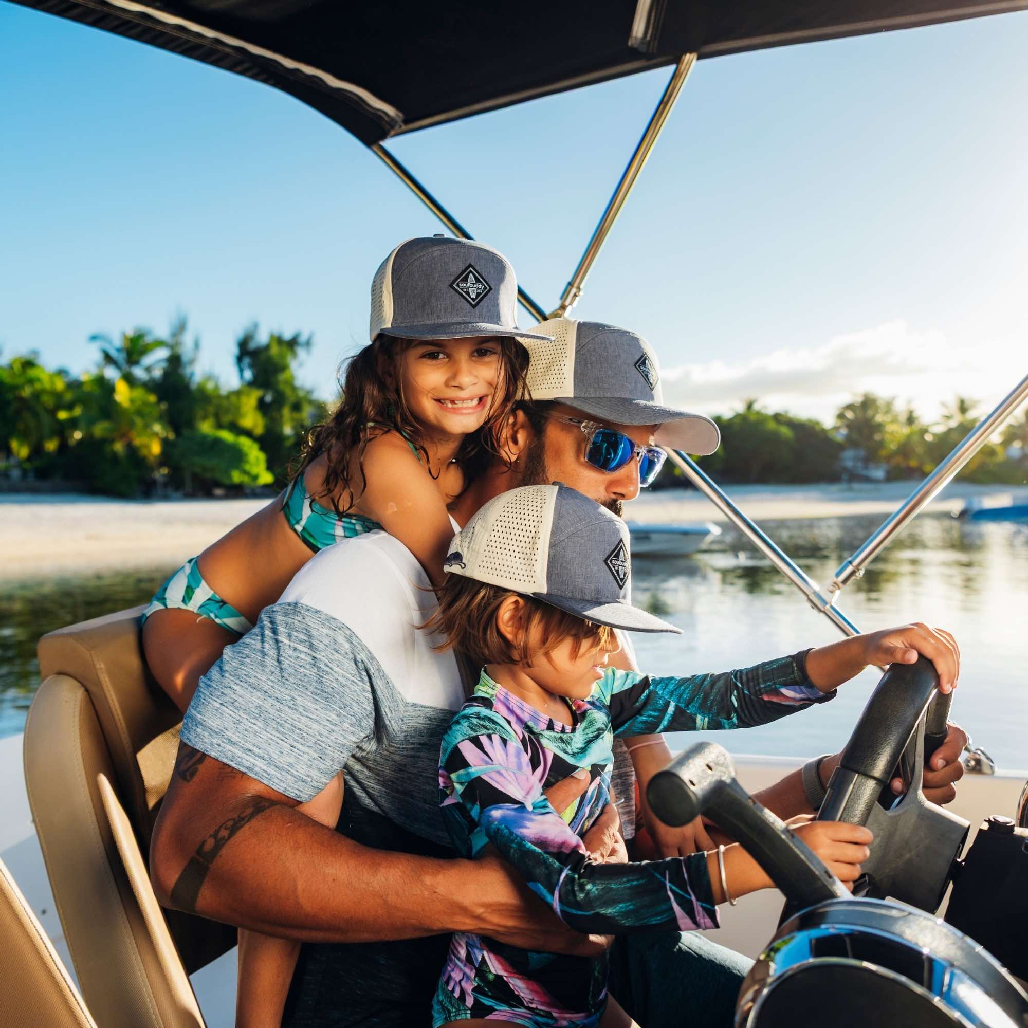 Vater mit zwei Kindern trägt graue Snapback Caps im Partnerlook auf einem Boot am Meer