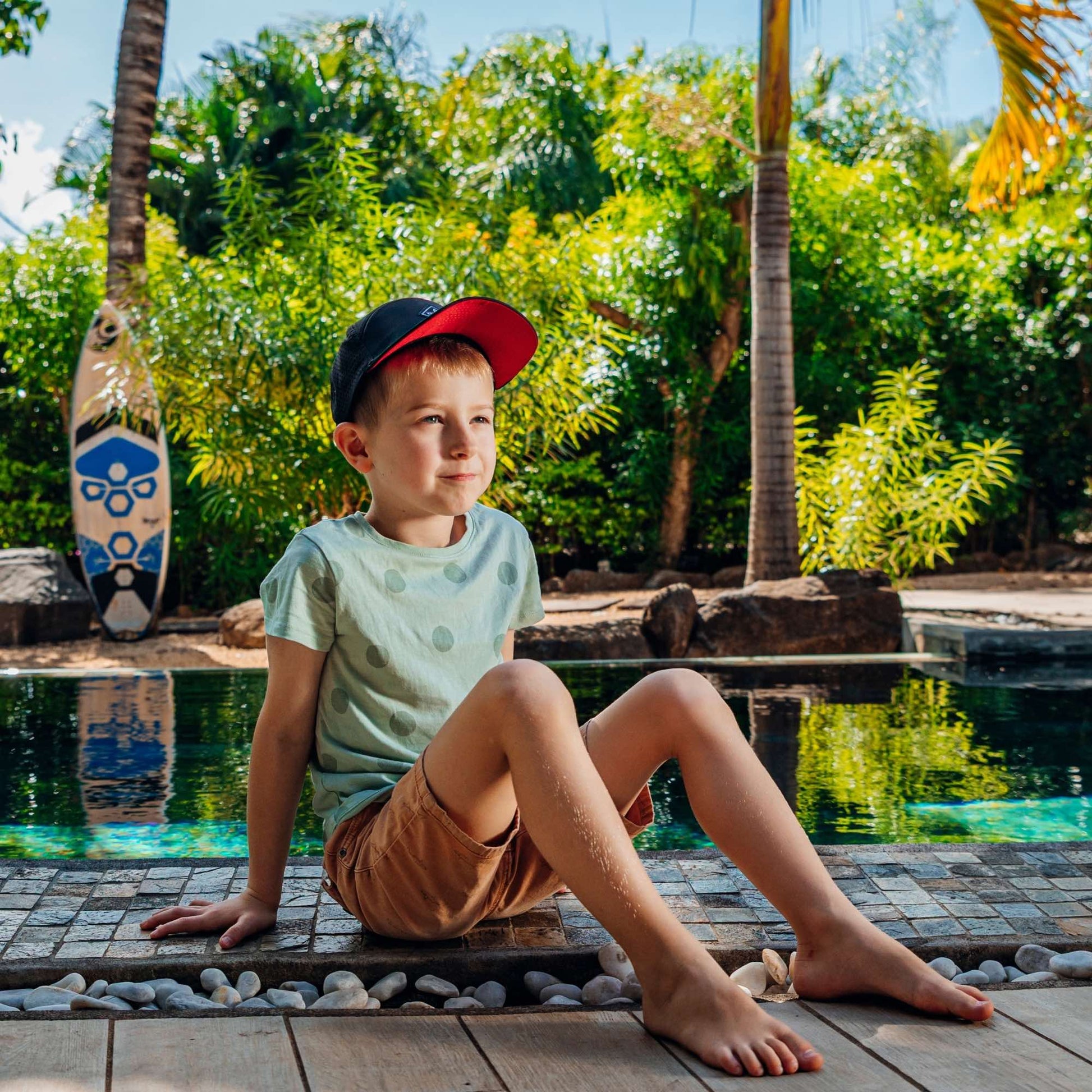 Kind mit schwarzer Snapback Cap sitzt entspannt am Pool in tropischer Umgebung bei sonnigem Sommerwetter