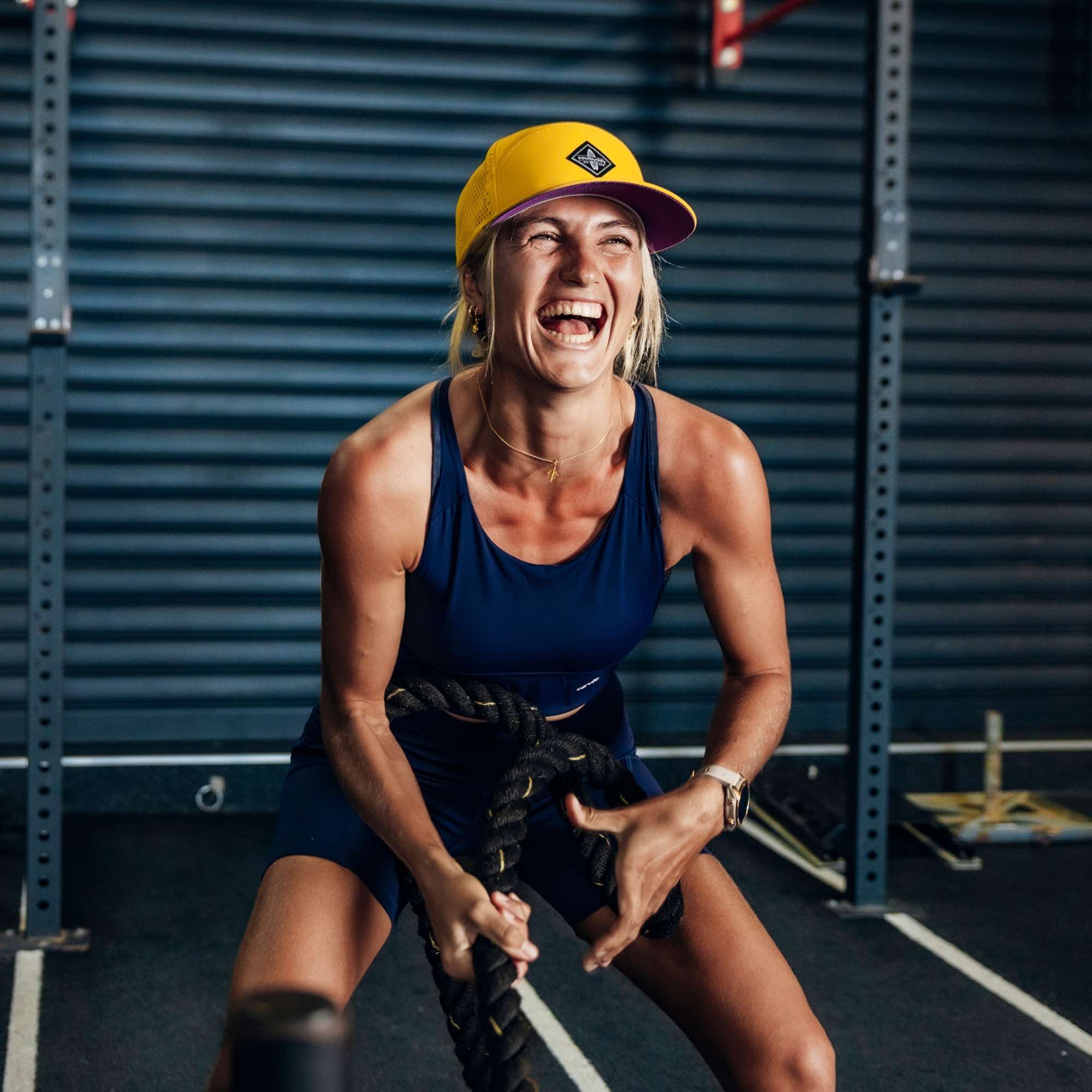 Sportliche Frau mit gelber Snapback Cap beim Battle-Rope-Training im Fitnessstudio, lacht und trainiert intensiv mit schweren Seilen