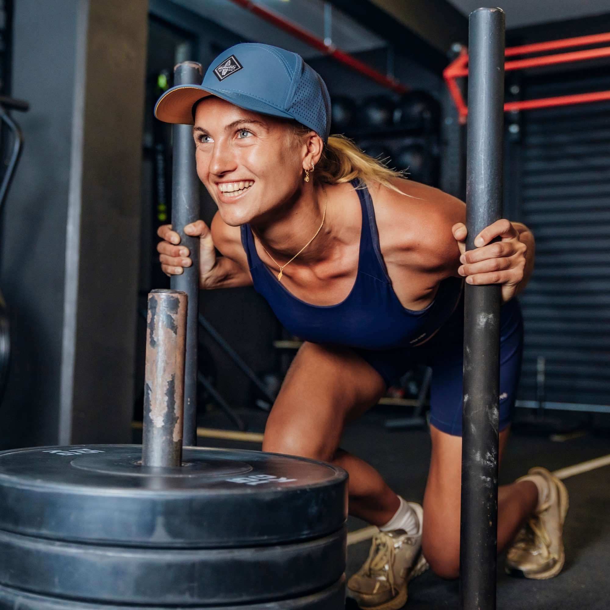 Sportliche Frau mit blauer Snapback Cap beim Sled Push im Fitnessstudio, schiebt einen Gewichtsschlitten und lächelt motiviert während des Trainings