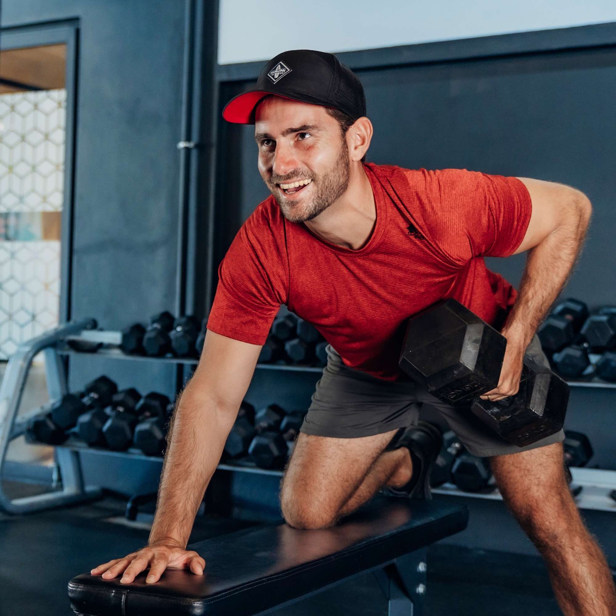 Sportlicher Mann mit schwarzer Snapback Cap und rotem Shirt beim Kurzhantel-Training im Fitnessstudio, führt ein einarmiges Rudern auf der Bank aus
