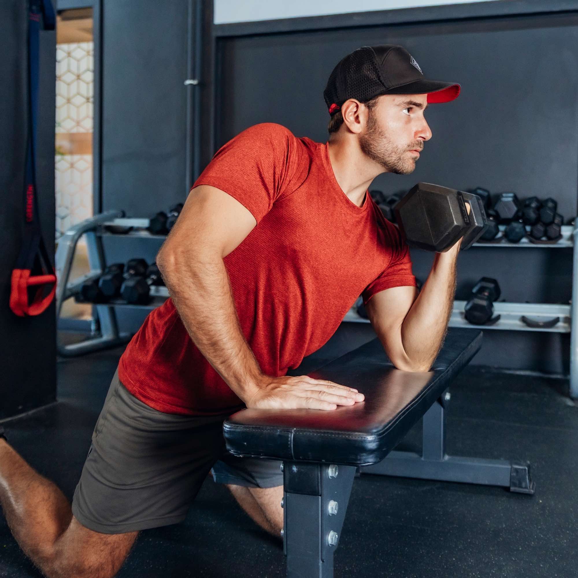 Sportlicher Mann mit schwarzer Cap und rotem Shirt beim Kurzhantel-Training im Fitnessstudio, stützt sich auf einer Bank ab und führt eine einarmige Bizepsübung aus