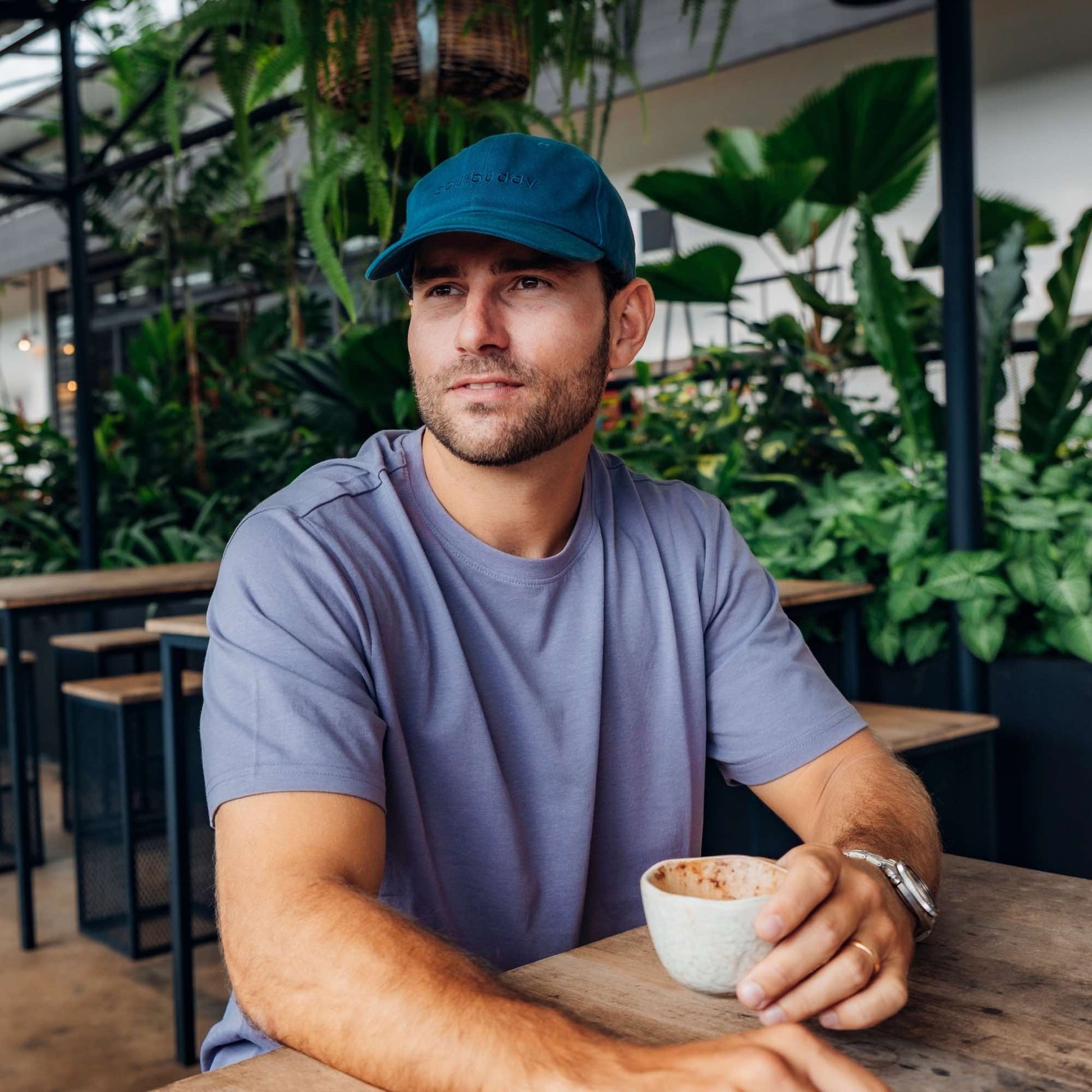 Mann mit petrolfarbener Soulbuddy Cap sitzt entspannt im Café und hält eine Tasse Kaffee in der Hand vor grüner Pflanzenkulisse