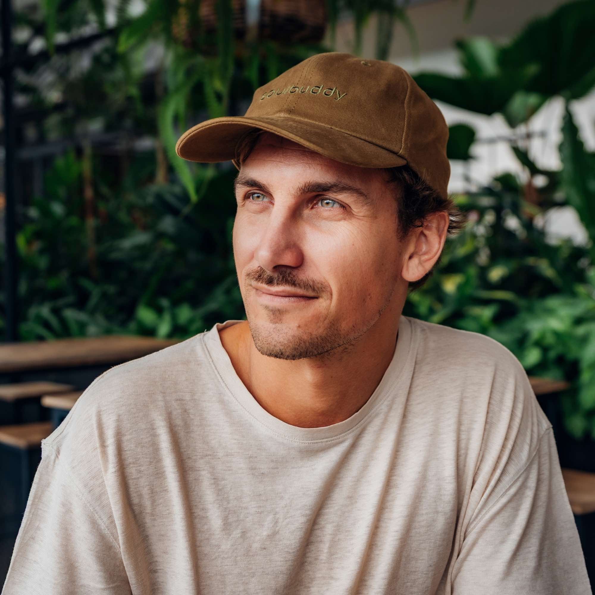 Mann mit olivfarbener Soulbuddy Cap blickt entspannt zur Seite in einer grünen, pflanzenreichen Café-Umgebung