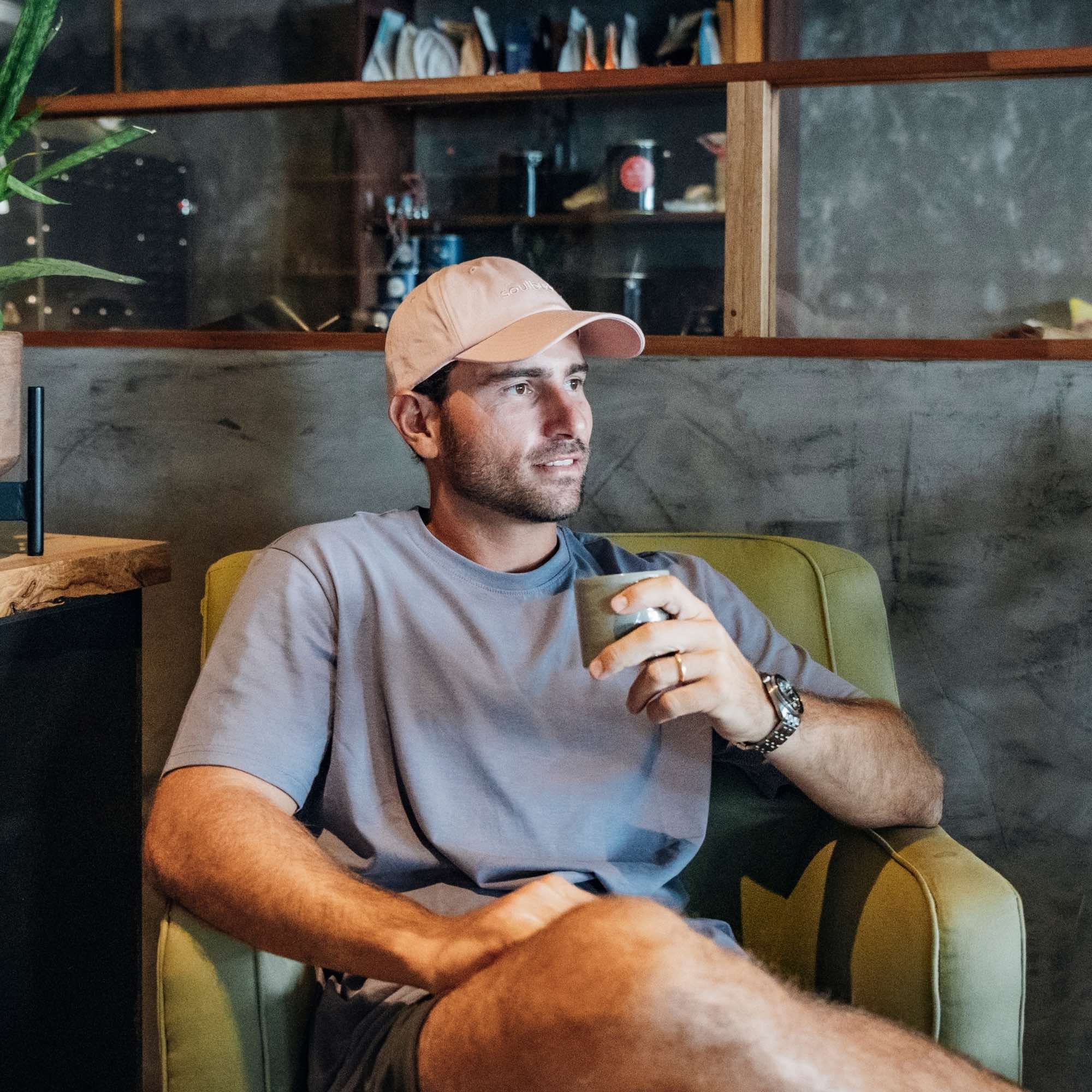 Mann mit creme-farbener Cap sitzt entspannt in einem Sessel und hält eine Tasse in der Hand in gemütlicher Café-Atmosphäre
