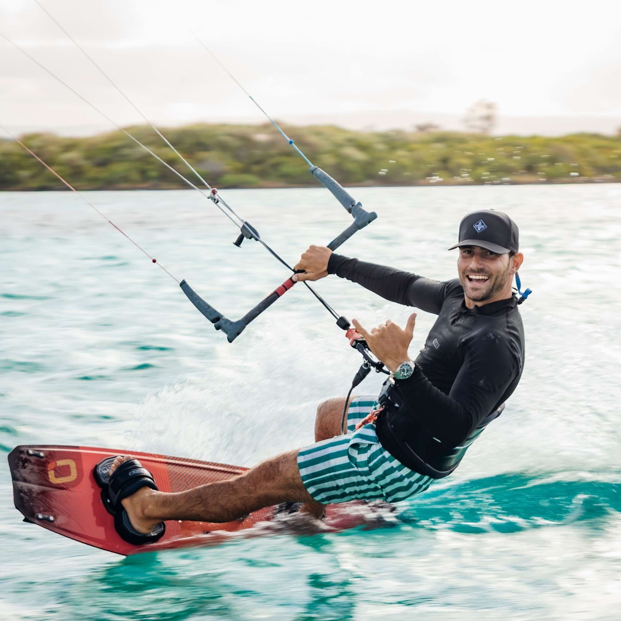 Sportlicher Mann mit schwarzer Snapback Cap beim Kitesurfen auf türkisfarbenem Wasser, macht Shaka-Handzeichen und lächelt in die Kamera