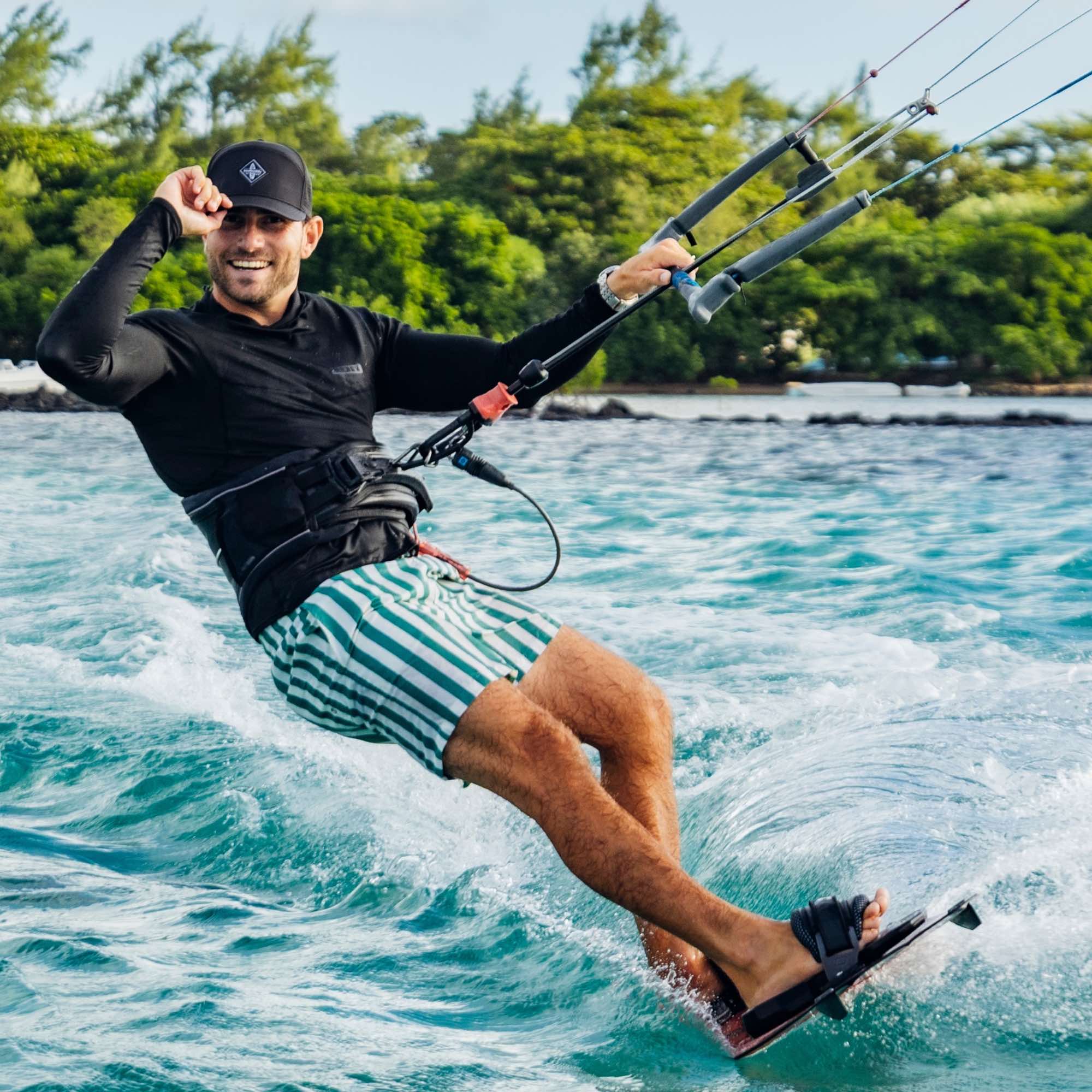 Sportlicher Mann mit schwarzer Cap beim Kitesurfen auf dem Meer, hält die Bar und zeigt ein Shaka-Handzeichen, während er über das Wasser gleitet