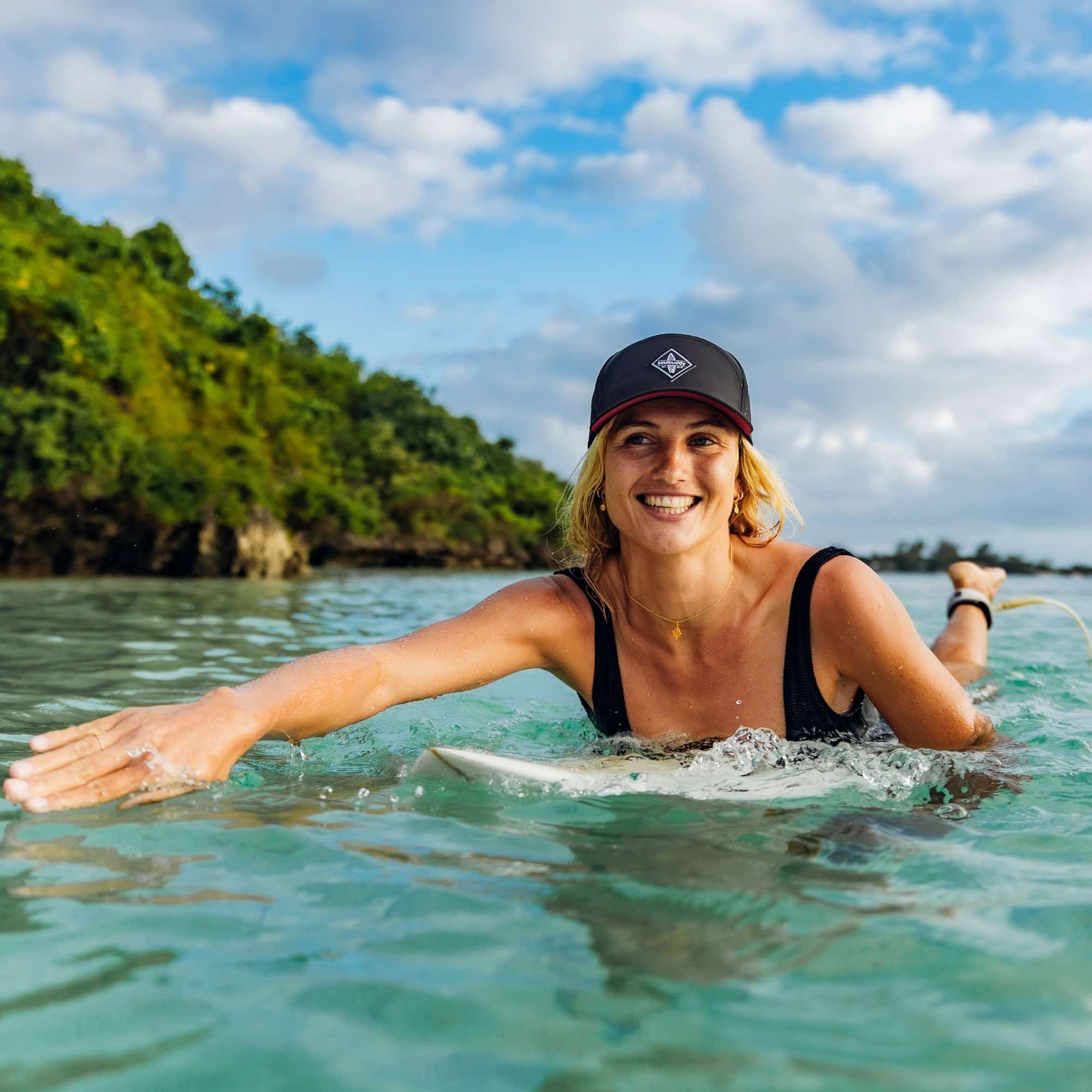 Sportliche Frau trägt schwarze Snapback Cap beim entspannten Surfen im Meer vor tropischer Küste bei sonnigem Wetter