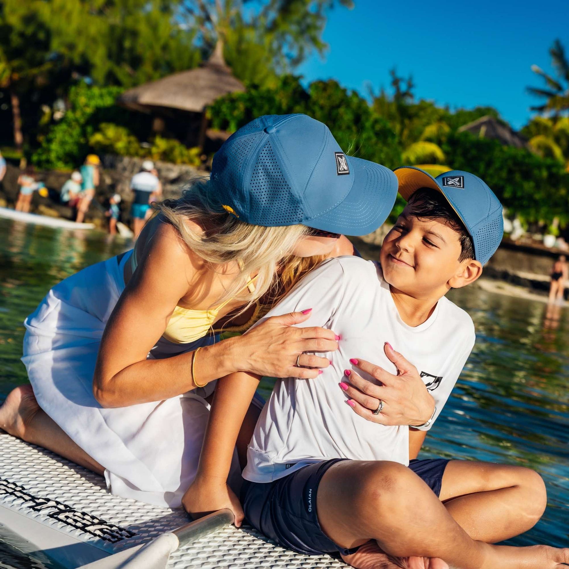 Mutter und Sohn mit blauen Snapback Caps im Partnerlook genießen liebevollen Moment auf einem Stand-Up-Paddle-Board im sommerlichen Urlaub am Meer