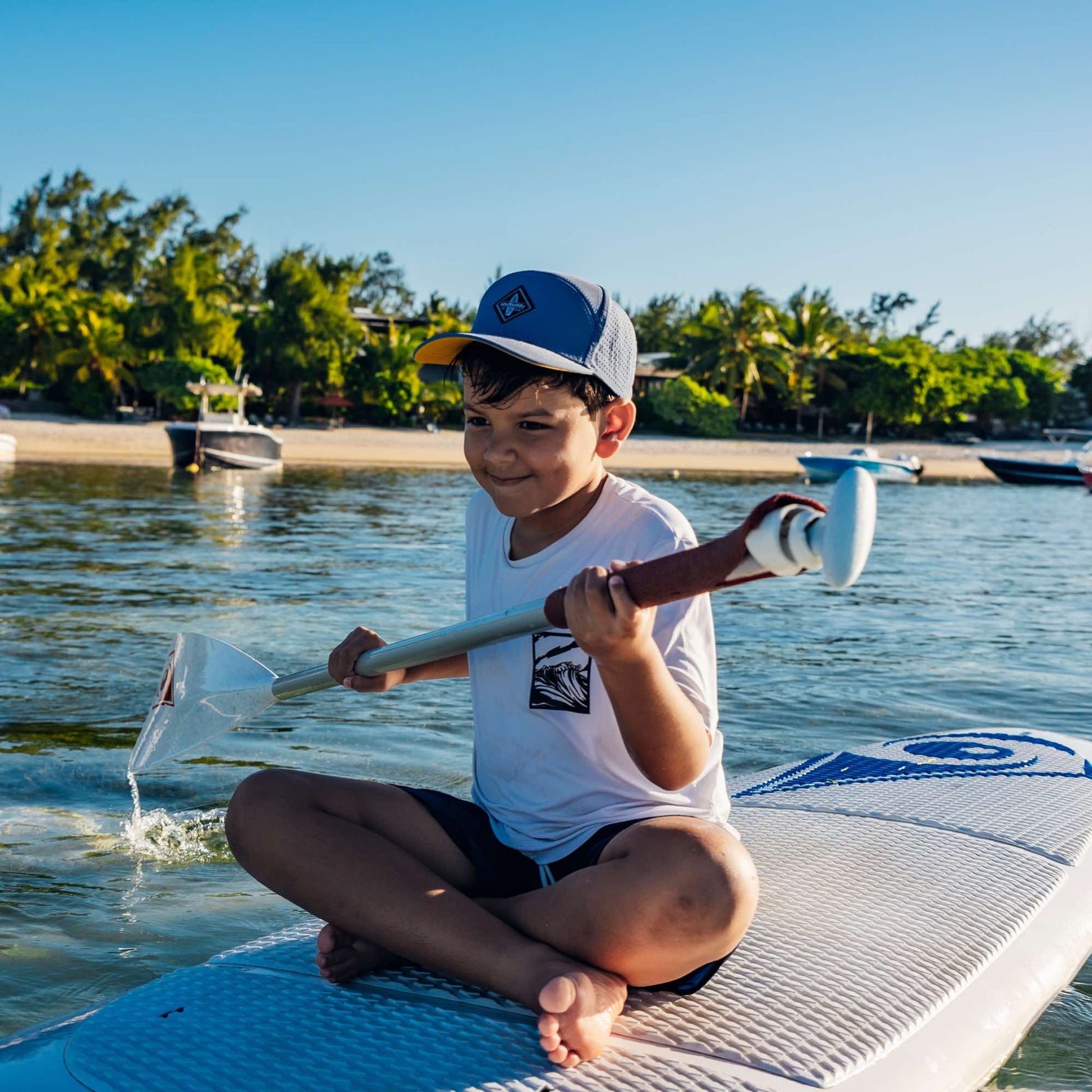 Kind mit grauer Snapback Cap paddelt auf einem Stand-Up-Paddle-Board im flachen Wasser am Strand bei sommerlichem Wetter